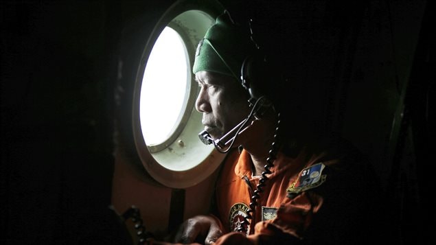 A crew member of an Indonesian Air Force C-130 airplane looks out of the window during a search operation for the missing AirAsia flight 8501 jetliner over the waters of Karimata Strait in Indonesia. It is a very dark photo. We see an Indonesian soldier with headphones and a microphone peering through a bright round window.