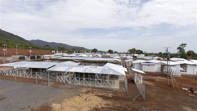 The Kerry Town Ebola treatment centre in Sierra Leone. We see a series of wood buildings with tin roofs sitting in a flat wilderness region. They sit on brown earth with small hills in the background.