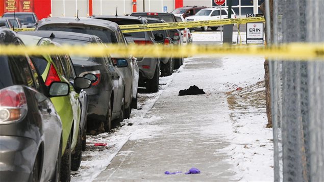 Discarded clothing lies on a Calgary sidewalk near where police are investigating a multiple shooting. We see a sidewalk half covered with snow along with cars parked at the curb on the left. Behind a yellow police tape, we see an unidentifiable piece of blue clothing in the foreground. Further down the sidewalk we see a pile of black clothes and more yellow police tape.