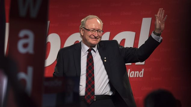 Wade MacLauchlan waves to the crowd as he is acclaimed leader at the Prince Edward Island Liberal leadership convention in Charlottetown on Saturday. We see the new premier, a handsome man with grey hair, standing with a smile on his face. He is wearing glasses and is dressed in a dark suit with the jacket open revealing a dark red plaid tie. He is waving with his left hand. Behind him is a red and white Liberal Party poster.