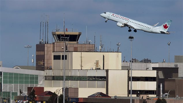 An Air Canada Jet taking off from Halifax Stanfield International Airport in Nova Scotia. The silver plane has her nose heading to the air. Below is the control tower and airport terminal.