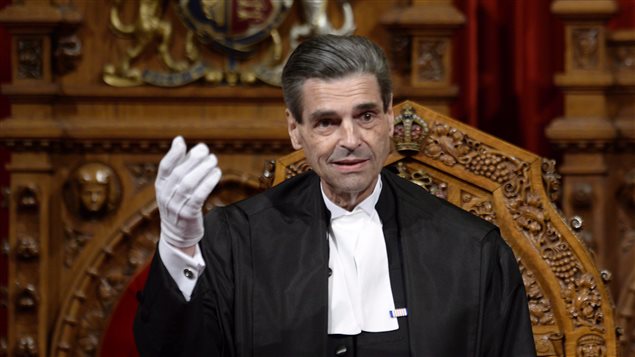 The death of Pierre Claude Nolin has left the Canadian Senate with another crisis to solve. We see Mr. Nolin in the Speaker's Chair. He is gesturing with his right hand, which is dressed in a white glove. He is wearing a dark robe and white ascot. He is thin with neatly combed black hair and dark eyes and appears to be looking out into the Senate Chamber.