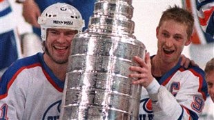 Edmonton's Wayne Gretzky, right, and Mark Messier hold up the Stanley Cup trophy, May 26, 1988 following their 6-3 win over the Boston Bruins in the finals. It was the Oilers' four championship in five years. We see Messier on the left, still wearing his helmet, and Gretzky (with short cropped hair) holding the Stanley Cup between them. Both are wearing enormous smiles and appear very, very young.