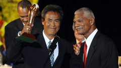 Former Expo Dennis Martinez, of Nicaragua, and former Expo manager Felipe Alou, right, during his induction into the Latin Baseball Hall of Fame during a ceremony in La Romana, Dominican Republic in 2010. Martinez, a handsome and dark-haired man is raising a golden trophy with a tight smile on his face while Mr. Alou has a gentle smile on his face. Both are standing behind a microphone wearing fancy suits.