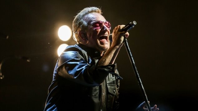 We see Bono in the foreground with his right hand on a mike. He is on stage with stage lighting in the background. He is wearing a leather jacket and his famed, big glasses. He exudes passion.