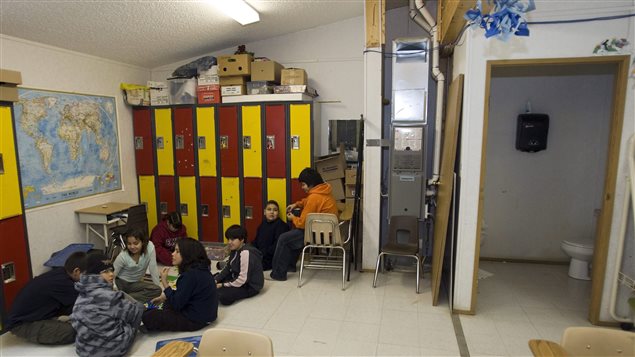 Report says more must be done to close the socio-economic gaps betwen aboriginals and non-aboriginals. We see eight children of elementary school age playing on the floor of a run-down classroom. They play in front of yellow and red lockers with cartons placed atop that run mostly to the ceiling. An exposed pipe rises to the right. A  support board to the right is only half painted. The door to the bathroom has been removed and sits against the wall next to bathroom. On a wall above the children is a large map of the world.