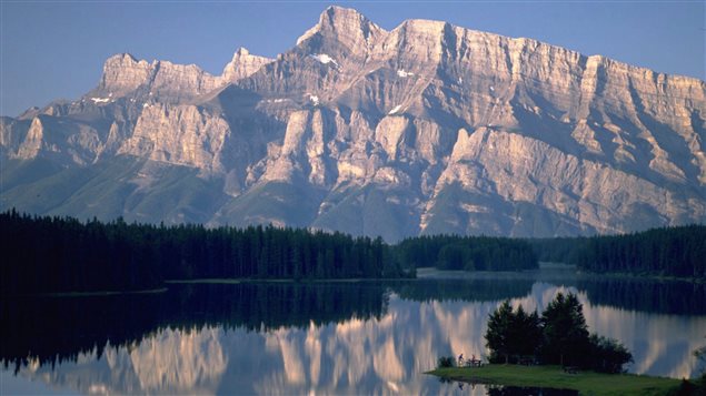 A view of Two Jack Lake in Banff National Park. Environmental groups fear Parks Canada is rushing guidelines that would result in too much development at a ski hill in the park. We see a wide shot of an absolutely gorgeous, peaceful and blue lake surrounded by deep-green pines. Rising above the lake and trees are the massive gray and snowcovered Rocky Mountains, which are reflected in the lake. Overhead, the sky is a azure.