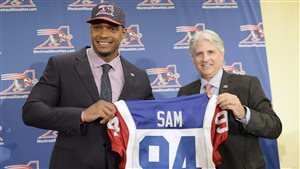 Sam and Alouettes General Manager Jim Popp were all smiles in May when Sam signed. Sam looking spiffy in his fancy suit and tie, and Popp, who is smaller, has white hair and is wearing a brown suit are holding in front of them (Sam is on the left) Sam's red, white and blue Alouettes jersey marked with the word 