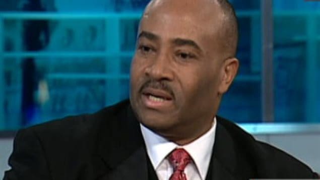 Senator Don Meredith joins three others tossed from the Tory caucus. We see a handsome African-Canadian man dressed in a dark suit, white dress shirt and dark tie.His head is shaved and he appears to be answering a question.
