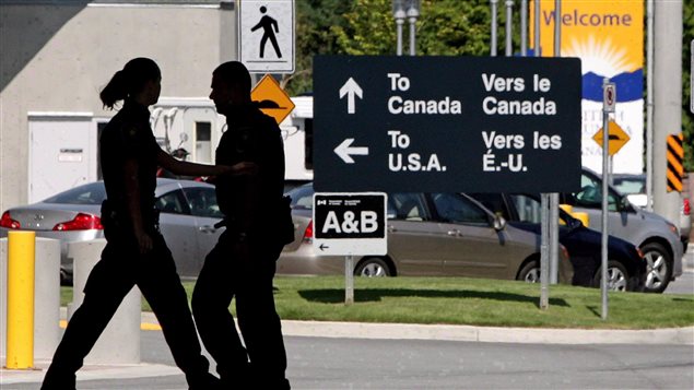 A Senate committee wants more light shined on the actions of Canadian border guards. We see (in silhouette) a female border guard on the left passing a male guard on the right. In the background we see a line of cars behind a large black sign with white letters and arrows that says "To Canada, Vers Le Canada" and "To U.S.A., Vers les É.-U."