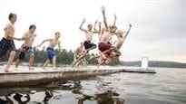 One of those joyful moments that makes our summers. We see seven or eight male teenagers leaping off a dock together into a grey-coloured lake.