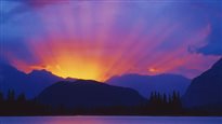 A summer dawn in the Canadian Rockies. We see the bright, yellow sun rising (with sunbeams) rising behind blue and purple mountains.