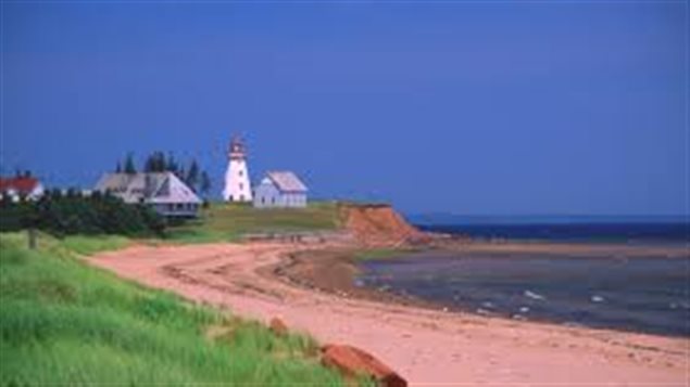 A summer's day on Canada's East Coast provides food for the sould. We see a long, pink beach with blue water to its right and a lighthouse and barn off in the distance. The sky is a deep blue.
