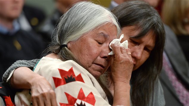 Residential school survivor Lorna Standingready is comforted by a fellow survivor in the audience during the closing ceremony of the Indian Residential Schools Truth and Reconciliation Commission in Ottawa earlier this month. We see an elderly, grey-haired lady using her right hand to wipe away a tear from her left eye. Her hair is partially braided and she is wearing a mainly white dress with a red, white and black star-shaped symbol on the right sleeve. She is being comforted by a dark lady who has her right arm around the older lady's shoulder.