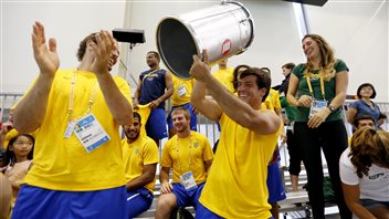 Supporters of the Brazil men's water polo team celebrate after the team scored a goal against Canada in Brazil's 11-9 victory on Tuesday, three days before the opening ceremonies. We see about a dozen young men and one woman--most dressed in yellow shirts--lapping and cheering. A man in the right is holding aloft what appears to be a small, silver kettle drum. All have wide smiles on their faces.