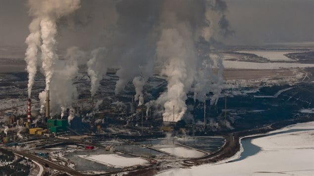 Fort McMurray, Alberta, the epicentre of oil extraction in Canada. We see an aerial shot (very wide) of factories belching streams of smoke into the atmosphere