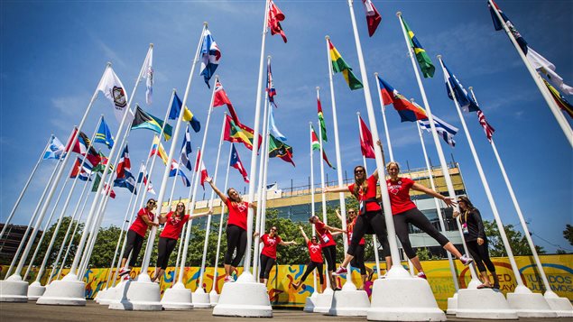 The Canadian women's water polo team poses for a picture on the flag poles inside the Pan American Games athletes village. Over 6,000 athletes are taking part in the Games. We see the girls dressed in red curling around the white poles holding the flags of the participating countries.