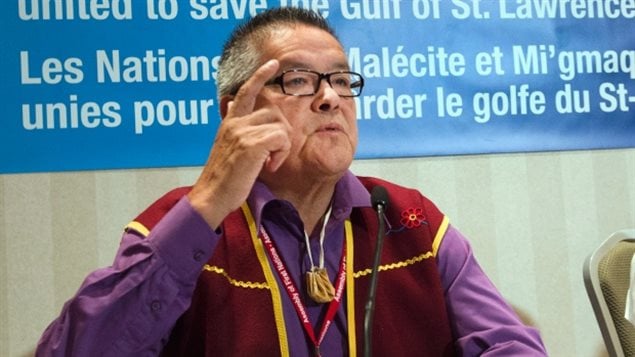 Innu Chief Jean Charles Pietacho speaks to reporters at the Assembly of First Nations congress on Wednesday in Montreal. The chief has short salt-and-pepper hair and is wearing horn-rimmed glasses. He is seated at a table and making a point by raising his right hand in the air. He is wearing a red vest with a touch of yellow embroidy over a blue shirt and has a tiny gold sack hanging from his neck.