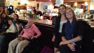 The launch earlier this year of the campaign leading to the adoption of a framework law in Quebec in terms of accessibility. Isabelle Ducharme is on the far left. We see four women, two of them in wheelchairs, in front of a softly-lit bar with the female bartender in the background. The glasses in overhead racks have light gleaming from them. In the background to the right of the four women in front are other customers, mostly women.