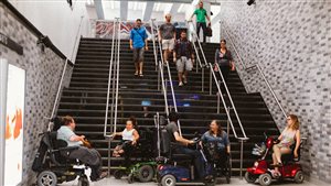 A Manifestation d'Accessibilize Montreal demo at the Place des Arts metro station in Montreal on July 25, 2014. More and more, people with disabilities are increasing their political activities. We see five people in motorized wheelchairs gathered at bottom of a four rows of stairs as five people heading to the trains begin the descend the stairs. The walls of the station are handsomely tiled. The stairwell and floor above are brightly lit.