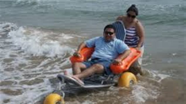 People in wheelchairs aren't asking for special favours. They just want public policies that allow them a shot at the kind of lives the rest of us enjoy, full access to public transportation, for example. We see a man in a wheelchair coming out of a lake being ashore pushed by a woman. It is a sparkling summer day, the water is blue-green. He is wearing a blue shirt and shorts. The woman is a white shirt with blue stripes and wearing white summer pants. Both the man and the woman are wearing sunglasses. The wheels of his chair are a bright yellow. 