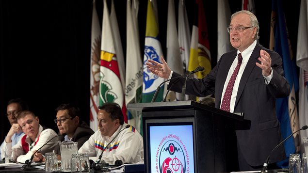 Former prime minister Paul Martin addresses the Assembly of First Nations congress in Montreal. Mr. Martin is standing behind a lectern with his arms spread wide as he makes a point. He hair is grey and he wears glasses. He is wearing a dark suit, red tie and white dress shirt. To his right at a head table, four chiefs listen. Behind them are the colourful flags of First Nations tribes. In the front of the lectern is the AFN symbol, a round circle filled with red animals. Attached to the circle and flowing downward are seven feathers.