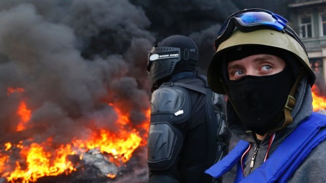 Anti-government protesters fleeing riot police in Kyiv camped in the main lobby of the Canadian embassy for at least a week during an uprising in January, 2014. We see two men in the foreground at the right of the picture. The one on the farthest right is wearing a grey battle helmet with purple goggles perched on top. He also wears a black mask that covers the lower part of his face. To his right, we see the back of another man, wearing an all-black battle suit, similar to someting from Star Wars. In the background, we see a yellow fire blazing with plumes of dark, grey smoke rising from it.