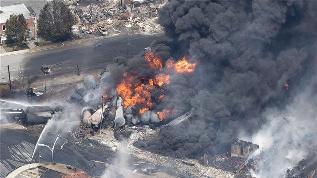 Smoke rises from railway cars that were carrying crude oil after derailing in downtown Lac-Megantic, on July 6, 2013. We see plumes of dark smoke flowing up from the blond blaze of a fire. We also see dozens of derailed tanker cars curled together and the ruins of the town in the background.