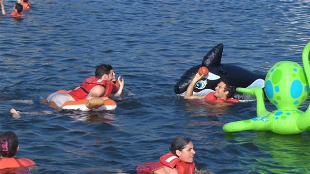 Swimmers enjoy life at Montreal's Old Port under a summer sun, but be careful what you wish for. We see young people swimming with plastic floats disguised as a yellow octopus and a black shark. The water is blue-grey and peaceful. 