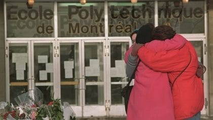 Three women embrace after laying flowers in front of École Polytechnique​ in Montreal, where a gunman killed 14 women on Dec. 6, 1989. All three are standing at the right of the picture. All are wearing red winter coats. In the background is the school building itself. Across its windows is written of École Polytechnique​ de Montreal. In the front of the building to the left, flowers have been laid.