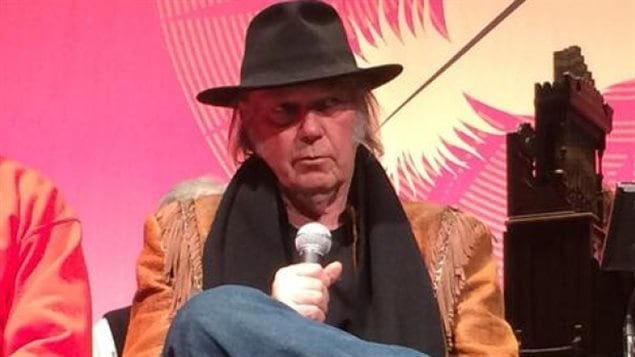 Neil Young is very unhappy with the effects of audio streaming. We see Young from the waste up seated with a microphone in his right hand. He is wearing a dark cowboy hat, a fringed buckskin jacket and has abrown scarf around his neck. He appears to be looking at someone who is asking him a question. In the background is a giant Indian feather surrounded by a pink background.
