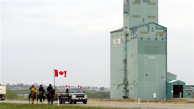 Riders and a support vehicle travel past a grain elevator on the Canadian prairies.