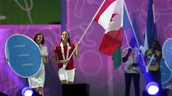 Kia Nurse, who led Canada to a gold medal in women's basketball, carries in the Canadian flag during Sunday's closing ceremonies. The picture is textured in blue and and pink colours. Nurse is wearing a red shirt and white pants and has the flag in front of her. To her right is a women model dressed in a white dress holding up an oblong sign with Canada's name spelled out three times (there's an accent over the final a in the 