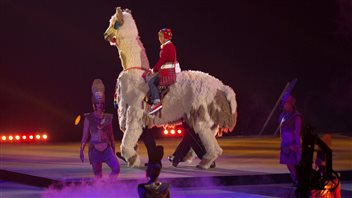 Performers stopped at nothing to celebrate at the closing ceremonies of Pan Am Games. We see a rider dressed in red perched atop of a giant (fake) llama. Rider and llama are surrounded by several other performers over a dark blue-lit floor.