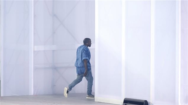Kanye West walks off stage at the Pan Am closing closing ceremonies on Sunday.  We see from a distance a tall man dressed mainly in a full-blown blue shirt and blue jeans in full stride about to walk off stage. He is dwarfed by giant blue and white wall.