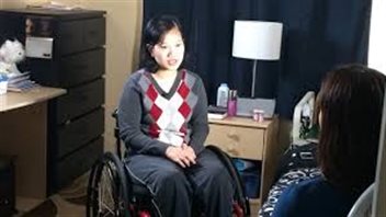 Vicky Venancio will receive the health care she needs to continue her rehabilitation. We see Ms. Venancio sitting in a wheelchair being interviewed in the aparment where she was in the picture at the top.  She is wearing the grey and white sweater knitted in diamond shapes we sas in the previous photo along with dark grey pants. She has short dark hair, an open face and is listening intently.