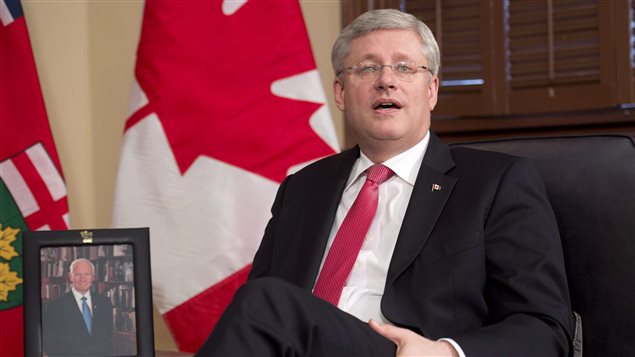 We see Mr. Harper sitting in a plush dark chair wearing a charcoal grey suit, white shirt and red tie. His legs are crossed and he is looking up with his mount partially open, making a point. He wears a tiny Canadian flag pin in his left lapel. To his right is a photograph of Governor General David Johnston. Behind Mr. Harper is a large Canadian flag.
