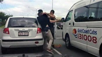 No surprise here: Road rage doesn't just affect Canadians. Here we see a couple of angry Australians. We see two men fighting at the rear of a silver compact car. The man on the right appears to be bare-chested and is attempting to strangle the other man who is reeling backwards. On the right is a white minibus on which the words Gold Coast Cabs is written in blue. Below the words are the numbers 131008.