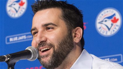 Blue Jays general manager Alex Anthopoulos finally had reason to smile after management loosened the purse strings and let him hit the trade market with full force. We see Anthopoulos sitting at a table behind a microphone with a big smile on his face. He has short black hair and an growing dark beard. Behind him on the wall are Blue Jays logos