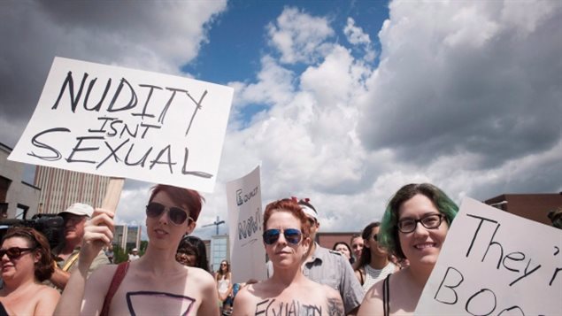 Some of the demonstrators who gathered Saturday in Waterloo, Ont., at the "Bare With Us" rally, organized to help raise awareness about women's right to go topless in Ontario. We see three young women face on from the shoulders up. Three are wearing sun glasses, two are holding placards. The girl second from left in the picture holds a placard stating "Nudity isn't Sexual." We only see part of the other placard. All we see are the words "They're."  The two girls in the middle have painted slogans on their chest, but we cannot read them. We see a crowd of men and women behind the four women in front. The blue sky is marked by dark, cumulus clouds.