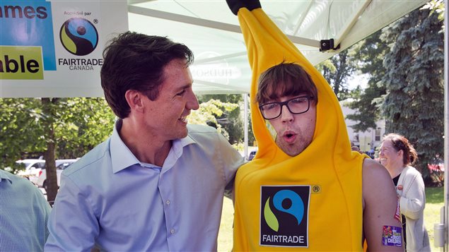 Liberal leader Justin Trudeau  greets a fair trade advocate at a music festival Saturday, in Hudson, Que. Trudeau moved his campaign to Toronto on Tuesday. We see Mr. Trudeau at the right of the photo dressed is a stylish blue Oxford shirt as he smiles and shakes the hand of a man dressed in what appears to be a yellow banana costume. At the top of the main part of the banana and below the stem, we see the man's face. He appears to be about 25 years old, has a early Beatles haircut and is wearing large horned-rim glasses. On the main part of the banana is a logo (somewhat resembling the yin-yang logo) on which is written "FAIRTRADE."