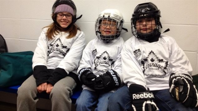 Kids at a Courage Canada hockey camp. They, too, now have a higher league to shoot for. We see three kids dressed in their white hockey seaters with a Courage Canada logo (a maple leaf and the words "Courage Canada). We have a tall girl with shoulder-length brown hair to the left. She is not wearing a mask. In the middle we have a short boy with a white birdcage helmet on.On the right a taller boy. He wears a dark birdcage helmet. The girl has a big smile. The boys are a bit more serious. This is hockey, after all.
