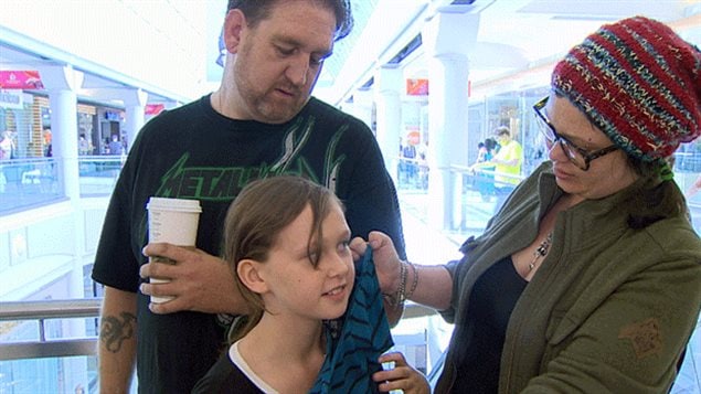 The falling Canadian dollar could be good news for retailers during the back-to-school shopping season. We see a family of three: the father in a black tee-shirt and holding a styrofoam cup of coffee in his left hand, stands behind his daughter who waits patiently as her mother, wearing glasses and a brown coat, lifts a shirt to the girl's face to see how it looks on her.