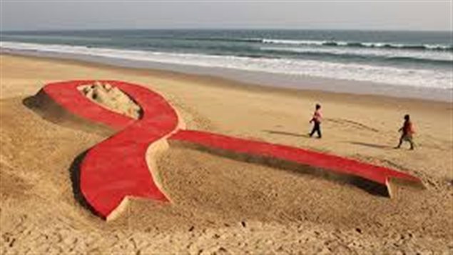  new study says early treatment and prevention is helping Canadians with HIV. We see two figures walking wide beach next to an ocean with the white surf coming in. On the beach is a giant red anti-AIDS ribbon symbol.