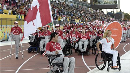 Team Canada, including Canada's flag-bearer Marco Dispaltro, centre, take part in the parade of nations during the opening ceremony at the Parapan American Games in Toronto on Friday. We see many athletes, some in wheelchairs, one on the left of the picture with one leg walking with crutches. They wear red jersies, grey pants and white caps  Some of the other athletes are walking behind them. Dispaltro is holding a very large Canadian flag.