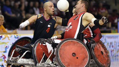 Colombia's Jhon Orozco Nunez, left, is rammed by Canada's Mike Whitehead as the ball comes loose during mixed gender wheelchair rugby matchwon 76-32 by Canada  on Sunday. We see two muscled men in large, red-wheeled wheelchairs converging with the  white ball being held in the hand of the man on the left, who has shaved head. He appears to be attempting a pass. His right arm, from the elbow down, does not exist. He wears a black jersey. His Canadian opponent has on a red jersey with a red maple leaf. A third man, barely visible, is coming in on his chair behind them.