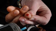 Type 2 diabetes is growing rapidly among South Asian men and black women in Canada. We see a closeup of a pair of hands, the left over the right. The ring finger of the bottom (right) hand has a spot of blood at its tip, the result of a diabetes test as view a small silver and black medical device in the foreground.