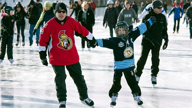 A father and son hit the ice. One wonders what's bubbling below the surface. We see a man in a dressed in a red Ottawa Senators hockey sweater holding his son's hand as they skate on a rink (likely the Rideau Canal). The boy is dressed in a black hockey uniform his left hand held in the air in joy. Behind them, we see lots and lots of other skaters in various forms of dress.