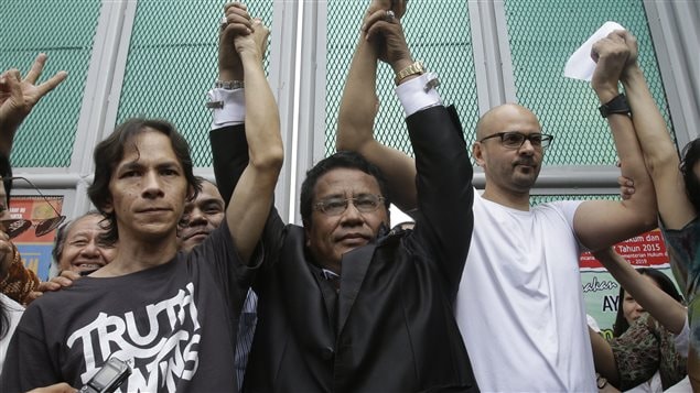 Neil Bantleman, right, his Indonesian teaching assistant, Ferdinant Tjiong, left, and their lawyer, Hotman Paris Hutapea, centre, celebrate their releases from prison on Friday. All three men have their raised hands clasped with one another  in celebration. However, their expressions are poker faced. Bantleman is a tall man with a shaved (or bald) head. He wears glasses and is dressed in a white tee-shirt. Tjiong is a shorter man. His dark hair runs down to his ears. He wears a dark tee-shirt with the words "Truth Wins" printed in white semi-cursive letters on the front. Hutapea wears rimless glasses and a dark suit that is crumbled as he raises his hands in victory.