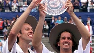 Daniel Nestor, left, and Sebastien Lareau of Canada hoist the trophy after winning the doubles final of the Tennis Masters Series tournament in Toronto in 2000, the same year they won the Gold Medal at the Beijing Olympics. Both are dressed in white and are wearing some kind of brown sombreros. Both have wide smiles on their face. Nestor is has long blond sideburns. Lareau has long hair and a moustache. He is holding the plate-trophy aloft with both hands as Nestor reaches to touch it with his right hand.